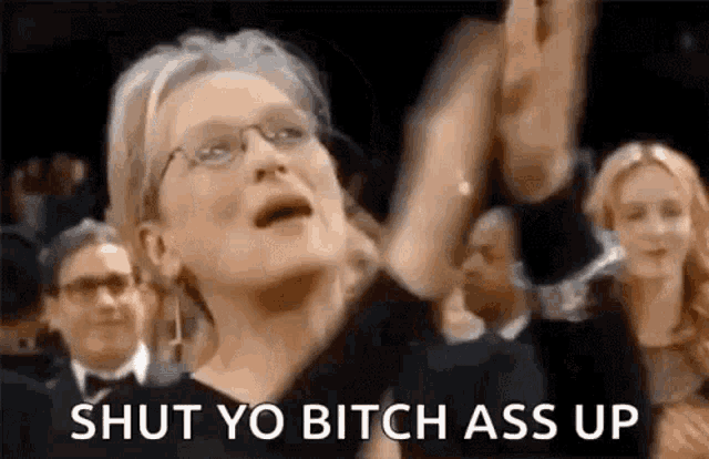 a woman wearing glasses is standing in front of a crowd with her hands in the air and says `` shut yo bitch ass up ''