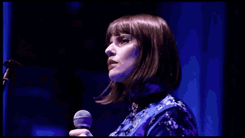 a woman in a blue dress stands in front of a microphone in a dark room