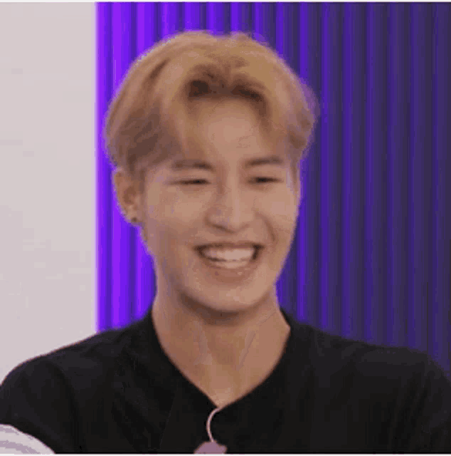 a young man with blonde hair is smiling in front of a purple curtain while wearing a black shirt .