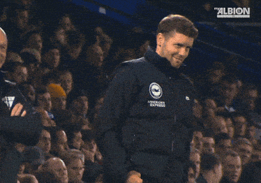 a man wearing an american express jacket gives a thumbs up in front of a crowd