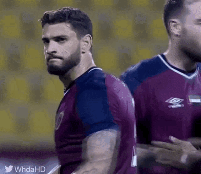 a man with a beard is standing next to another man wearing a purple shirt with the word umbro on it