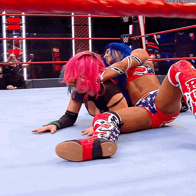 a female wrestler with pink hair is laying on the floor while another wrestler holds her