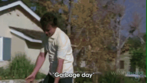 a man in a white shirt is standing in front of a house and saying garbage day