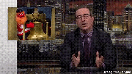 a man in a suit and tie is talking in front of a picture of a puppet and a bell