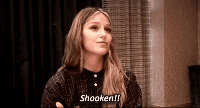 a woman in a plaid shirt is sitting in front of a curtain and saying shooken .