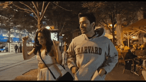 a man wearing a harvard sweatshirt walks down a street with a woman