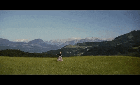 a woman in a long dress is standing in a grassy field with mountains in the background