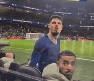 a man in a blue shirt is standing on a soccer field while another man looks on .