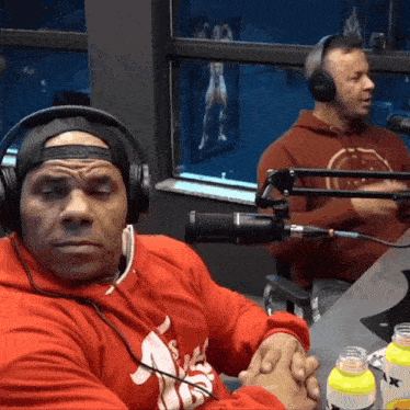 a man wearing headphones sits in front of a microphone while another man wearing a red hoodie sits behind him