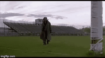a man in a trench coat is walking on a field next to a tree .
