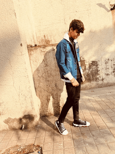 a young man wearing a denim jacket and black pants is walking on a sidewalk