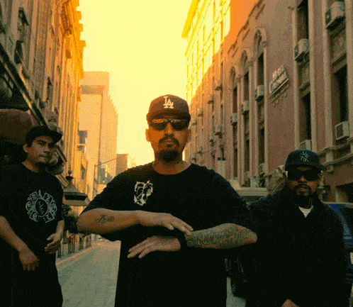 a man wearing a la hat stands with two other men on a city street