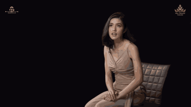 a woman in a tan dress sits in a chair in front of a black background that says tpm