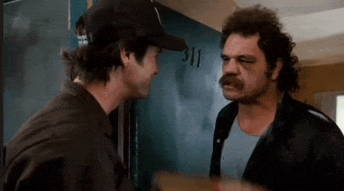 two men are standing next to each other in front of a door . one of the men is wearing a hat .