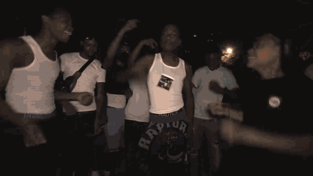 a group of people dancing in a dark room with a man wearing a white tank top