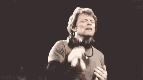 a man with a beard is standing in front of a microphone and clapping his hands in the air .