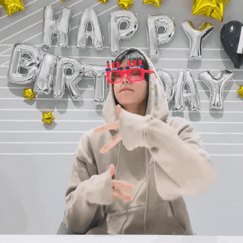 a man wearing a hoodie and sunglasses stands in front of balloons that spell happy birthday
