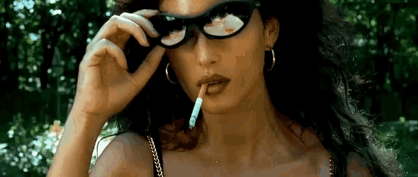 a woman wearing sunglasses is smoking a cigarette in a park .