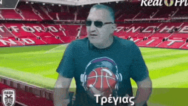 a man wearing sunglasses and a t-shirt with a basketball on it stands in front of a soccer field .
