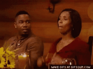 a man and a woman are sitting at a table talking to each other . the woman is pointing at the man .