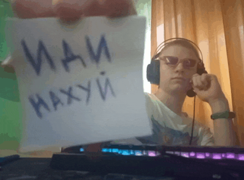 a man wearing headphones and sunglasses is holding a piece of paper that says ' иди нахуй ' on it