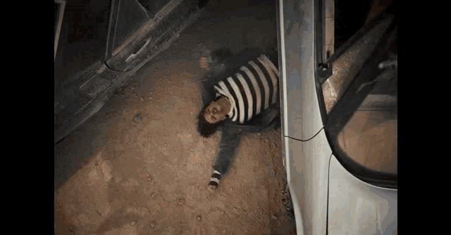 a person in a striped shirt laying on the ground next to a car