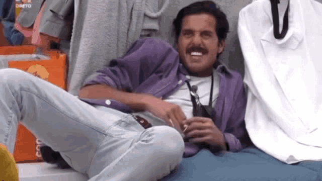 a man in a purple shirt sits on a bed with his legs crossed
