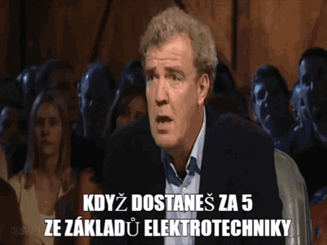 a man in a suit is sitting in front of a crowd and says kdyz dostanes za 5 ze základu elektrotechniky