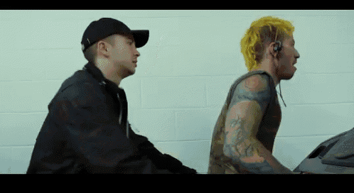 two men are standing next to each other in front of a white wall . one of the men has a yellow mohawk .