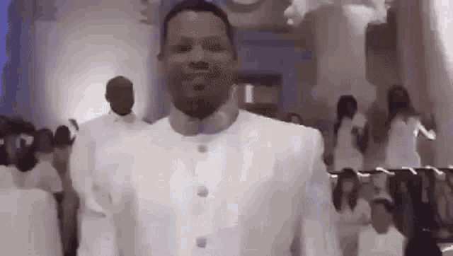a man in a white suit is standing in front of a crowd of people in a room .