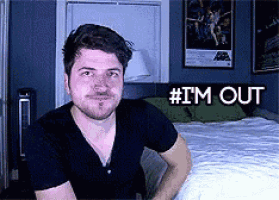 a man in a black shirt is sitting in front of a bed with the words # i 'm out above him