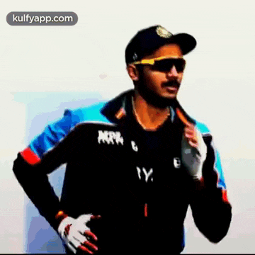 a man wearing sunglasses and a hat is standing with his hands on his hips and looking at the camera .