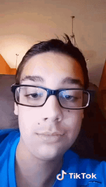 a young man wearing glasses and a blue shirt has a tiktok watermark on his shirt
