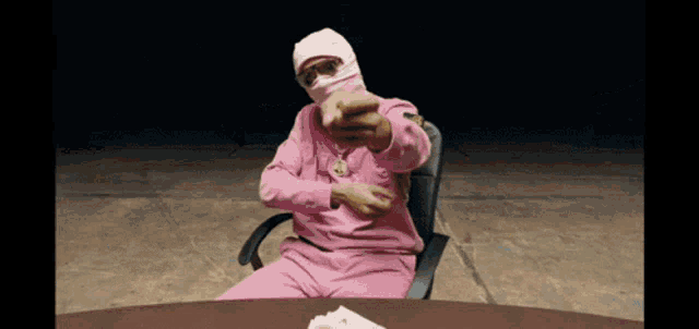 a man wearing a pink mask is sitting at a table and pointing at the camera
