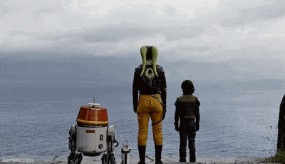 a woman and a boy are standing next to a robot on a ledge overlooking the ocean .