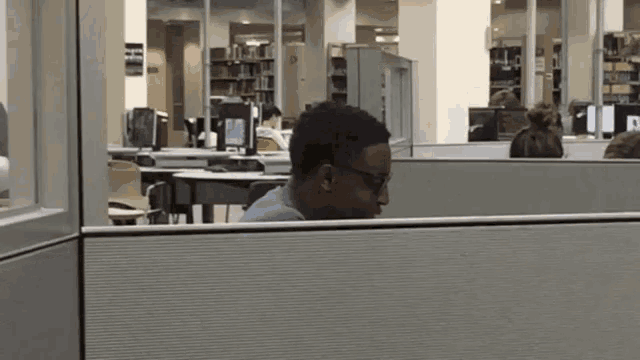 a man wearing glasses sits in a cubicle in front of a computer