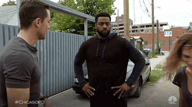 a man with his hands on his hips talks to another man and a woman in front of a nbc logo