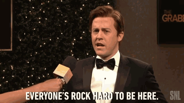 a man in a tuxedo says everyone 's rock hard to be here in front of a microphone