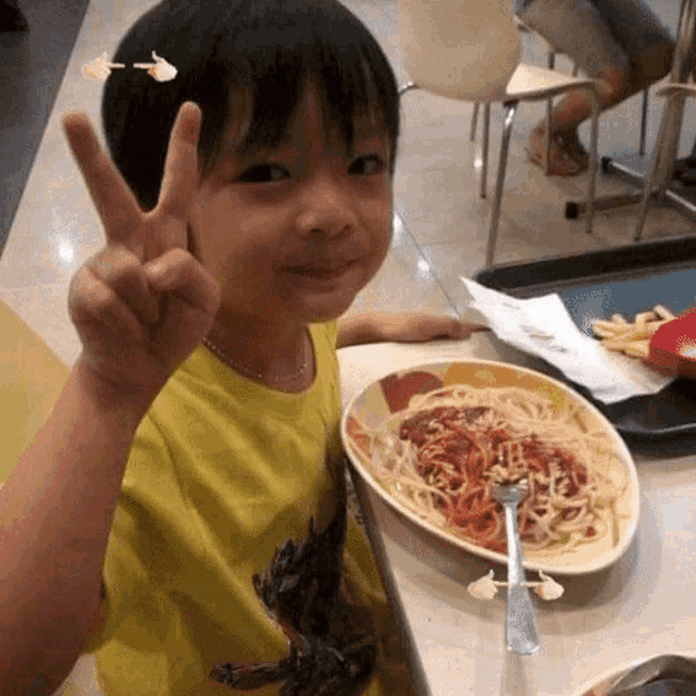 a young boy giving a peace sign in front of a plate of spaghetti and french fries