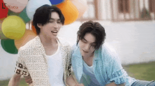 two young men are sitting next to each other in front of balloons and laughing .