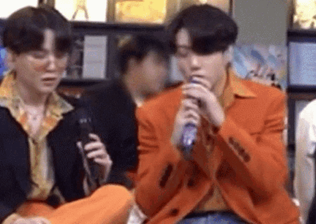 a man in an orange jacket is singing into a microphone while another man looks on .