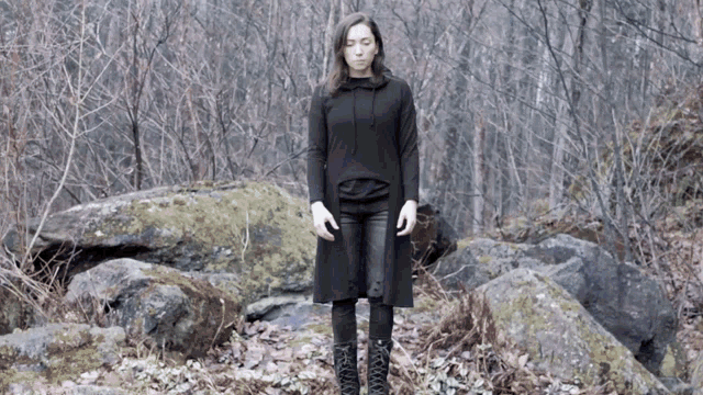 a woman in a black coat and black boots stands in the woods