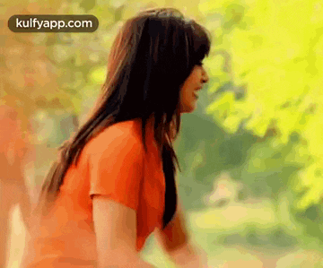 a woman in an orange shirt is standing in front of a tree in a park .