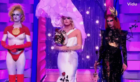 three drag queens are standing next to each other on a stage and one is wearing a bride 's dress .