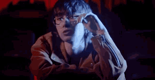 a man wearing glasses is sitting in a dark room with his hands on his face .