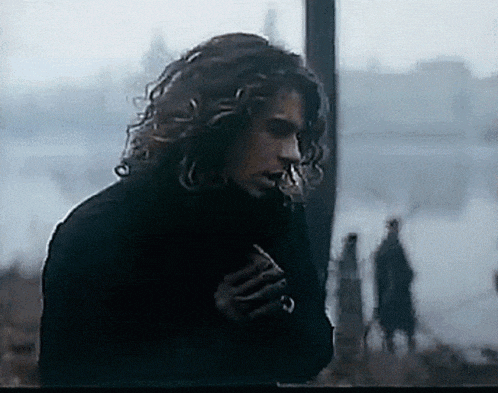 a man with long curly hair is smoking a cigarette while standing in the fog .