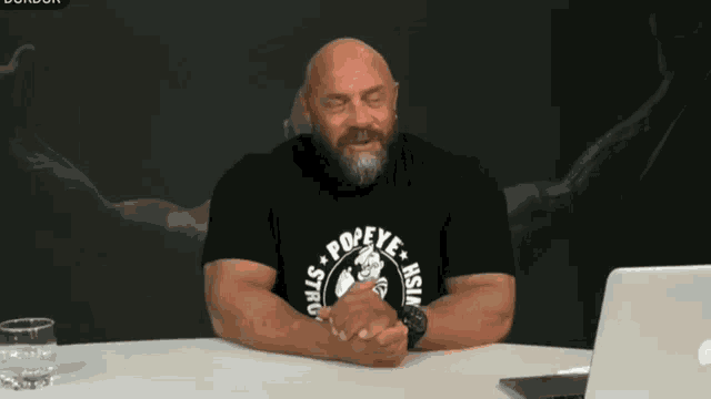 a bald man with a beard wears a black shirt that says popeye asia