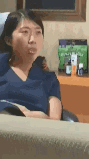 a woman in a blue scrub top is sitting on a couch in front of a table with bottles on it .