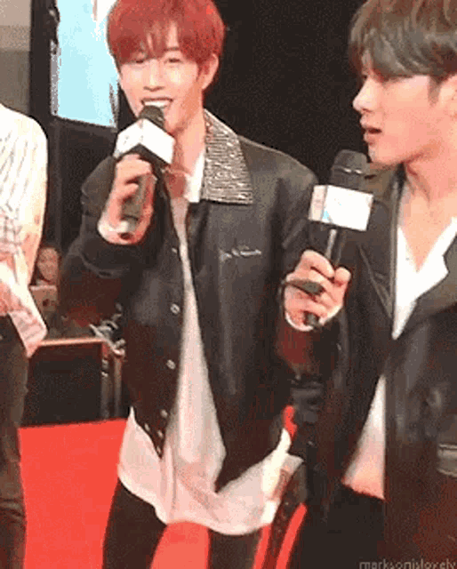 a man in a leather jacket is holding a microphone while standing next to another man on a red carpet .