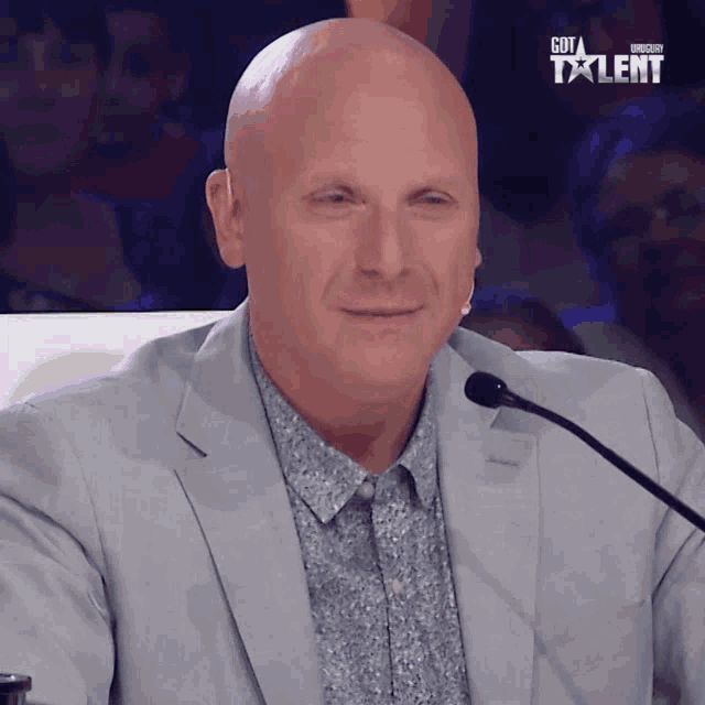 a bald man in a suit stands in front of a microphone with the words got talent on the bottom